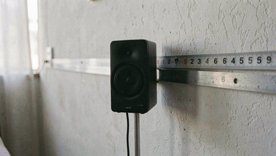Kompaktlautsprecher auf einem schmalen Metallständer, dahinter die Wand mit angelegtem Massband, das den Abstand zeigt, kühles Tageslicht von der Seite, halbnahe Aufnahme