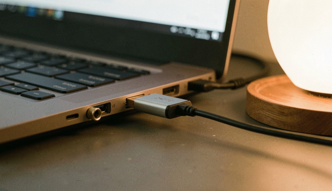 Kleiner USB-Adapter mit kurzem Kabel hängt am Anschluss eines Notebooks, daneben ein aufgeschraubter Kopfhöreranschluss, Bildschirmlicht und eine warme Lampe von rechts, Nahaufnahme